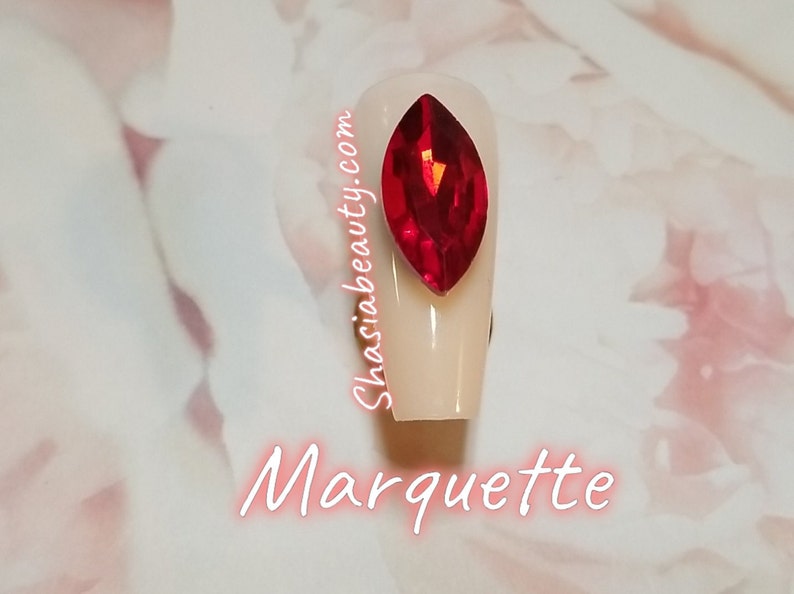 May include: A close-up of a white nail with a large, red, faceted, marquise-shaped rhinestone. The nail is surrounded by a white background with the text "Shasiabeauty.com" and "Marquette" written in pink.