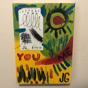 May include: A yellow, green, and red abstract painting with the words "You Me" and "JG" written on it. The painting also includes a small white square with the numbers "1 2 3 4 5 6 7" and the word "ella" written on it. The date "8-13-21" is also written on the white square.