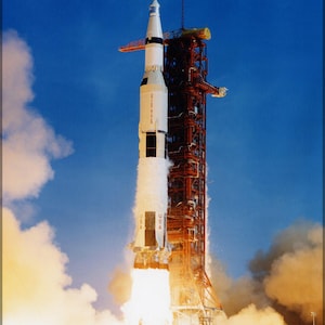 May include: A white and orange rocket with the words "United States" and "USA" on the side is launching from a launchpad. The rocket is surrounded by a large cloud of smoke and fire.