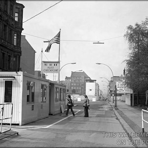 Könnte beinhalten: Ein Schwarzweißfoto eines Kontrollpunkts in Berlin, Deutschland. Das Foto zeigt ein Gebäude mit einem Schild, auf dem "Allied Checkpoint" steht. Im Hintergrund weht eine amerikanische Flagge. Zwei Personen gehen im Vordergrund.