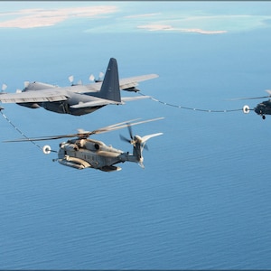 May include: A grey military cargo plane refuels a grey helicopter in mid-air over a blue ocean. A black helicopter is also being refueled by the cargo plane.