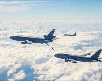 May include: Three military aircraft flying in formation over a field of white clouds. The aircraft are all gray and have the American flag on their tails.