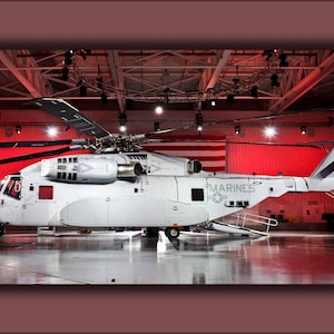 Pode incluir: Um helicóptero militar branco com a palavra "MARINES" no lado, estacionado em um hangar com paredes vermelhas e um teto de grelha metálica.