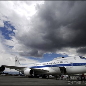 May include: A white airplane with blue accents and the words "United States of America" on the side. The plane is parked on a tarmac with a cloudy sky in the background.