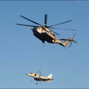May include: A large military helicopter is lifting a smaller white aircraft with a cable. The helicopter is flying against a clear blue sky.