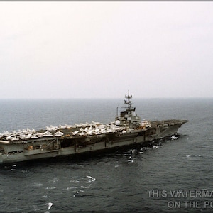 Könnte beinhalten: Ein grauer Flugzeugträger mit einer großen Anzahl von Kampfflugzeugen, die auf dem Flugdeck geparkt sind. Der Träger fährt auf einem ruhigen, grauen Meer.