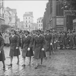 Puede incluir: Un grupo de mujeres con uniforme caminan por una calle de una ciudad. Todas llevan abrigos largos y sombreros. Las mujeres caminan en fila y todas miran hacia adelante.