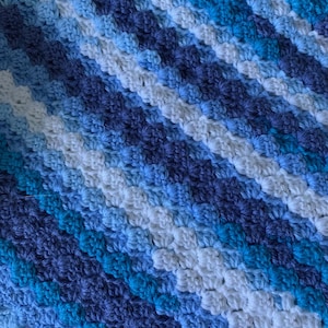 May include: A handmade crochet blanket featuring a diagonal pattern of varying shades of blue and white. The textured surface of the blanket is visible, showcasing the detailed stitch work. The color palette includes light blue, navy, and white.