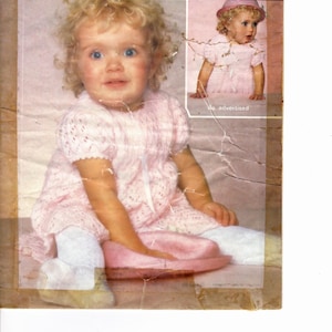 May include: Vintage baby knitting pattern featuring a young child in a pink knitted dress and matching hat. The pattern is for 4-ply yarn and includes sizes 21-23 inches. The Sirdar and Snuggly Baby Yarns logos are visible.