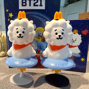 May include: Two RJ character figures from BT21, each featuring a white fluffy head, orange scarf, and yellow McDonald's arch crown, sitting on a light blue cratered base with a yellow flame, supported by a circular stand in white or black.