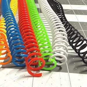 May include: A collection of colorful spiral binding coils in orange, yellow, blue, red, green, white, and black. These plastic coils are arranged in parallel rows, showcasing their vibrant hues and coiled structure. The coils are on a gray and white checkered surface.