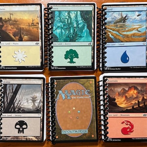 May include: A collection of six Magic: The Gathering card notebooks. Each notebook features a different landscape image, including plains, forest, island, swamp, and mountain scenes. The notebooks are bound with black spiral binding.