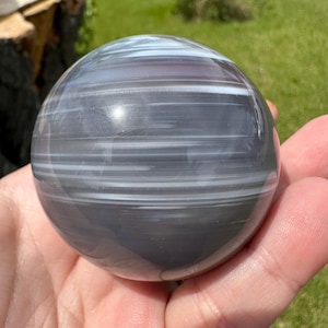 May include: A polished spherical stone featuring horizontal bands of dark gray and translucent white. The surface is smooth and reflective, displaying natural mineral layers that wrap around the orb in a consistent, linear pattern throughout the sphere.