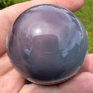 May include: A smooth, polished spherical stone with a deep, translucent purple and grey hue. The surface is highly reflective, showing a clear mirror-like finish that captures the surrounding environment in its rounded, glossy, and solid form.