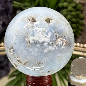 May include: A polished light blue chalcedony sphere featuring natural white inclusions and small vug cavities across its surface. The sphere is displayed on a dark red wooden stand next to a silver United States quarter coin for scale.