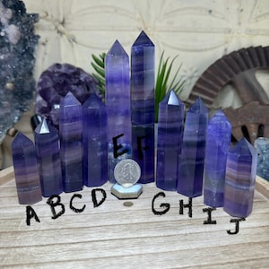 May include: A collection of ten purple fluorite crystal towers, each with a unique pattern and shape. The crystals are arranged on a wooden tray, labeled A through J. A coin is placed in front of the crystals for size comparison.