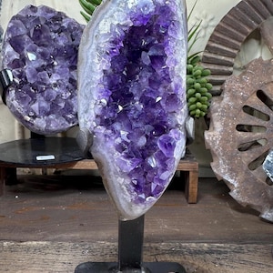 May include: A large amethyst geode crystal on a black metal stand. The geode features a deep purple interior with crystal formations and a white and gray outer edge. Another amethyst geode is visible in the background.