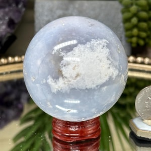 May include: A polished, light blue spherical stone featuring a prominent, textured white mineral inclusion across its surface. The sphere is mounted on a dark reddish-brown, tiered wooden stand, with a small silver coin visible for scale.