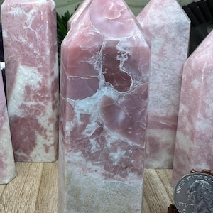 May include: Several pink gemstone obelisks with white veining are displayed. The obelisks have a rectangular base and a pointed top. The gemstones have a polished surface and are arranged on a wooden surface.