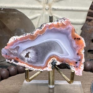 May include: A polished agate geode slice, showcasing bands of orange, white, and gray hues. The stone is mounted on a brass stand with a clear acrylic base, highlighting its natural patterns and textures. The geode features a central cavity.