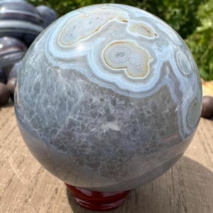 May include: A polished spherical stone featuring a smooth surface with intricate banding in shades of gray, white, and tan. The sphere displays a translucent crystalline texture in the lower section and sits on a small, dark red circular stand.