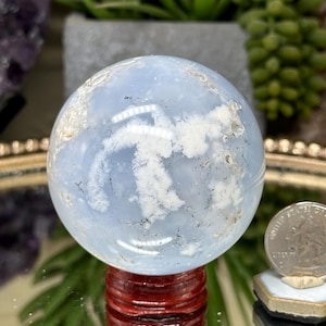 May include: A polished light blue sphere with white cloud-like inclusions and dark mineral veins, resting on a dark red wooden stand. A silver United States quarter coin is positioned to the right for scale, showing the size of the stone.