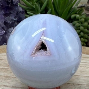 May include: A polished, light blue crystal sphere with a geode-like opening. The sphere sits on a small, round, reddish-brown wooden stand. The crystal has white and pinkish-purple veining. The background includes greenery and other crystals.