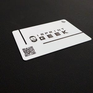 May include: A white card with the text "IMPRINT GEEK" in black, a logo of a bespectacled face, and a QR code. It also has a contactless payment symbol. The card is on a black textured background.