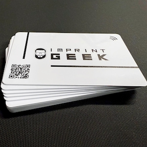 May include: A pile of white cards with rounded corners, displaying the text "IMPRINT GEEK" in black. A QR code and a stylized icon are also visible. The cards are stacked on a black surface.