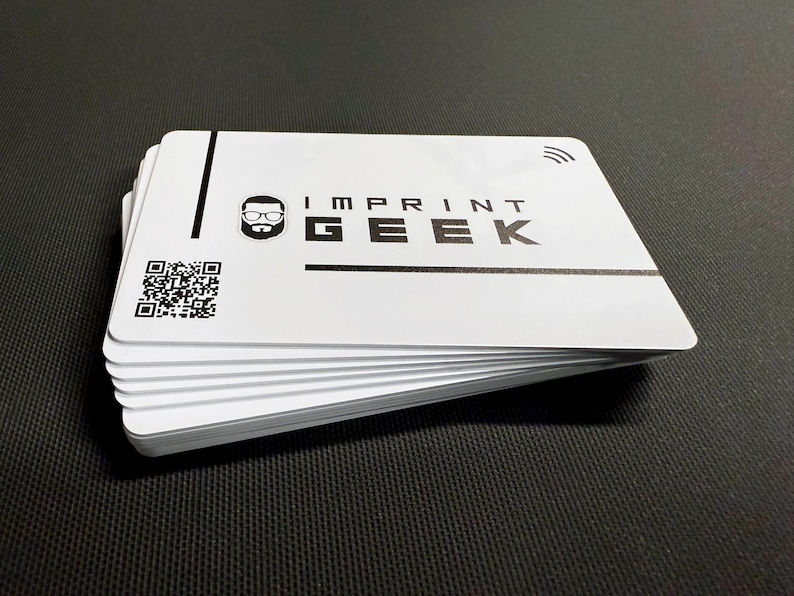 May include: A pile of white cards with rounded corners, displaying the text "IMPRINT GEEK" in black. A QR code and a stylized icon are also visible. The cards are stacked on a black surface.