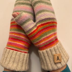 May include: A pair of colorful, striped fingerless gloves. The gloves feature a ribbed, cream-colored cuff and a multi-colored striped pattern in shades of red, orange, green, pink, and purple. A wooden tile with the letter "A" and the number "1" is attached to one cuff.