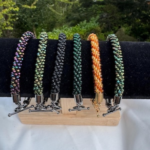 May include: Six beaded bracelets in various colors, including iridescent, green, black, and orange, are displayed on a black surface. Each bracelet features a silver-tone toggle clasp. The bracelets are presented on a wooden stand.