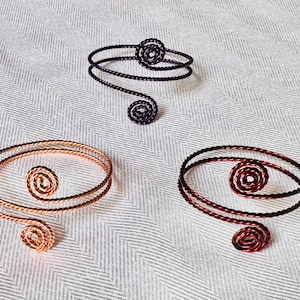 May include: Three arm cuffs in copper, dark purple, and burgundy. Each cuff features a spiral design at the top and bottom, with a circular spiral in the center. The cuffs are made of twisted wire.