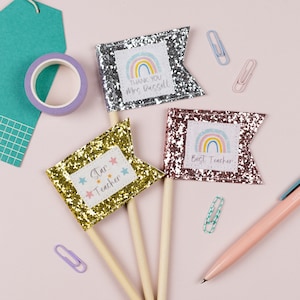 Puede incluir: Tres banderas de brillantina con diseños de arcoíris y estrellas. Las banderas dicen "Gracias Sra. Russell", "Maestro Estrella" y "Mejor Maestro".