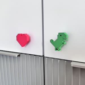 Puede incluir: Un imán pixelado en forma de corazón de color rosa brillante y un imán pixelado en forma de dinosaurio de color verde brillante sobre una superficie blanca. Ambos objetos presentan un diseño de bloques estilo 8 bits con acabado mate.