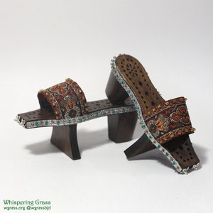 Op de afbeelding: Een paar bruine houten platform sandalen met ingewikkelde uitsnijdingen en een decoratieve stoffen band. De sandalen hebben een turquoise en zilveren kralen rand.