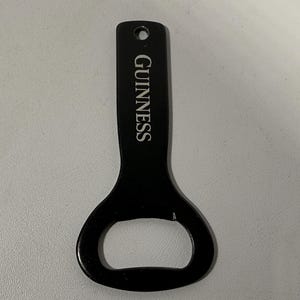 May include: Black bottle opener with the word "GUINNESS" printed vertically in white. The opener has a hole at the top for hanging and a rounded end for opening bottles. The opener is made of metal.