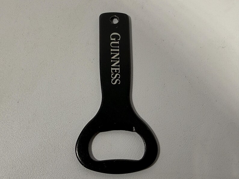 May include: Black bottle opener with the word "GUINNESS" printed vertically in white. The opener has a hole at the top for hanging and a rounded end for opening bottles. The opener is made of metal.