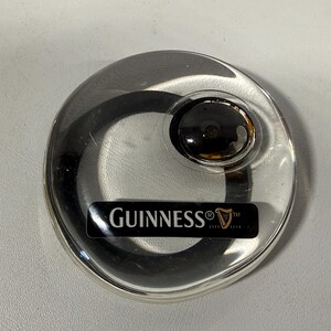 May include: A clear, circular Guinness coaster with a black and gold center. The coaster has a black rectangular label with the Guinness logo and text.