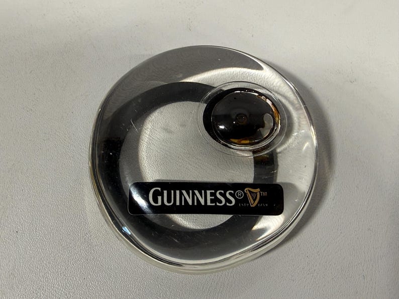 May include: A clear, circular Guinness coaster with a black and gold center. The coaster has a black rectangular label with the Guinness logo and text.