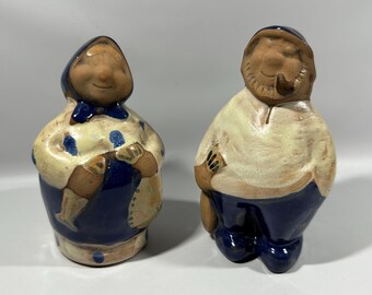 May include: A pair of ceramic figurines depicting a man and a woman. The woman is wearing a blue and white dress and a blue hat. The man is wearing a white shirt and blue pants. Both figurines have painted details and are in a rustic style.