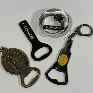 May include: Four bottle openers and a bottle stopper are displayed. One black bottle opener has "Guinness" printed on it. Another is shaped like a bottle with a keychain. A gold-colored opener has "Nitro IPA" printed on it. A silver bottle stopper has the Guinness logo.