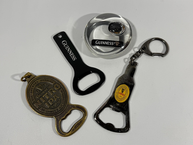 May include: Four bottle openers and a bottle stopper are displayed. One black bottle opener has "Guinness" printed on it. Another is shaped like a bottle with a keychain. A gold-colored opener has "Nitro IPA" printed on it. A silver bottle stopper has the Guinness logo.