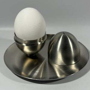 May include: A stainless steel egg cup with a white egg, a salt shaker, and a round plate. The egg cup holds the egg upright. The salt shaker is egg-shaped. The plate is round with a curved edge.
