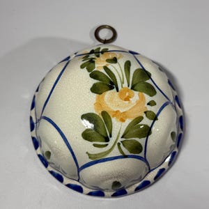 May include: A white ceramic mold with a floral design. The mold features a yellow and green flower in the center, surrounded by green leaves. Blue lines and dots decorate the edges. A small metal ring is attached at the top.