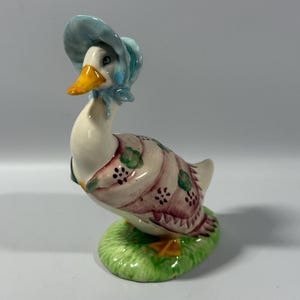 May include: A ceramic duck figurine with a light blue bonnet and a patterned dress. The duck has a yellow beak and feet, and is standing on a green base. The dress has pink, green, and black details.