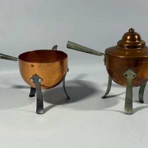 May include: Two copper fondue pots with metal handles and legs. One pot is open, the other has a lid with a small knob. The copper has a warm, reddish-brown hue, and the metal legs and handles are silver.