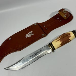 May include: A brown leather sheath with a silver eagle logo and a silver blade hunting knife with a brown and white stag handle. The knife has the text "Packards Silver Eagle" on the blade.