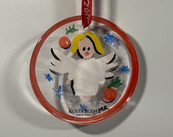 May include: Clear, round Christmas ornament with a red border, featuring a painted angel with yellow hair and white wings. The ornament has a red ribbon for hanging and the text "KOSTA BODA MR SINCE 1742".