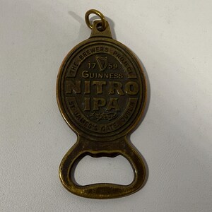 May include: A brass-colored bottle opener with a circular top featuring the text "NITRO IPA" and "GUINNESS" with the year "1759". The opener has a loop at the top and a bottle cap opener at the bottom.